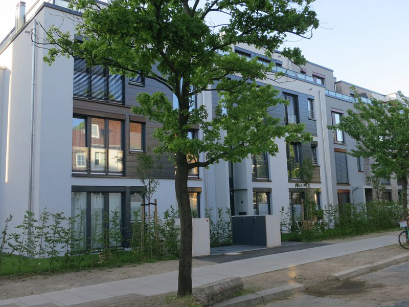 Ein moderner, kubischer Wohnungskomplex mit weissen Fassaden und großen Fensterfronten an einerm begrünten Gehweg mit jungen Büschen und zwei kleinen Laubbäumen in der Loehnstraße Hannover
